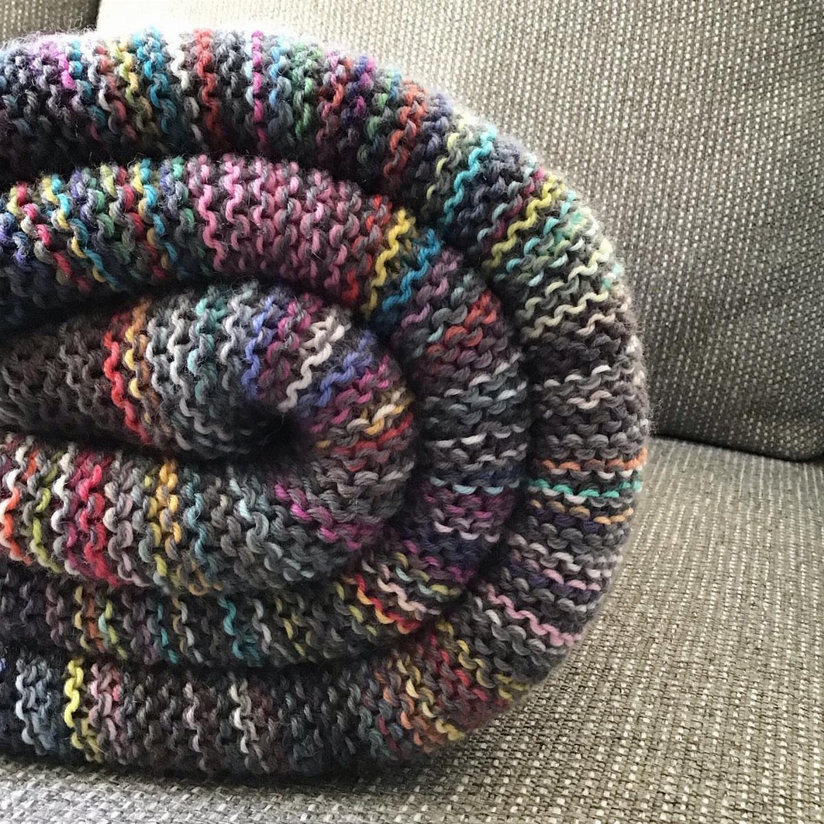 Rolled multicolored chunky knit blanket resting on a grey textured sofa.