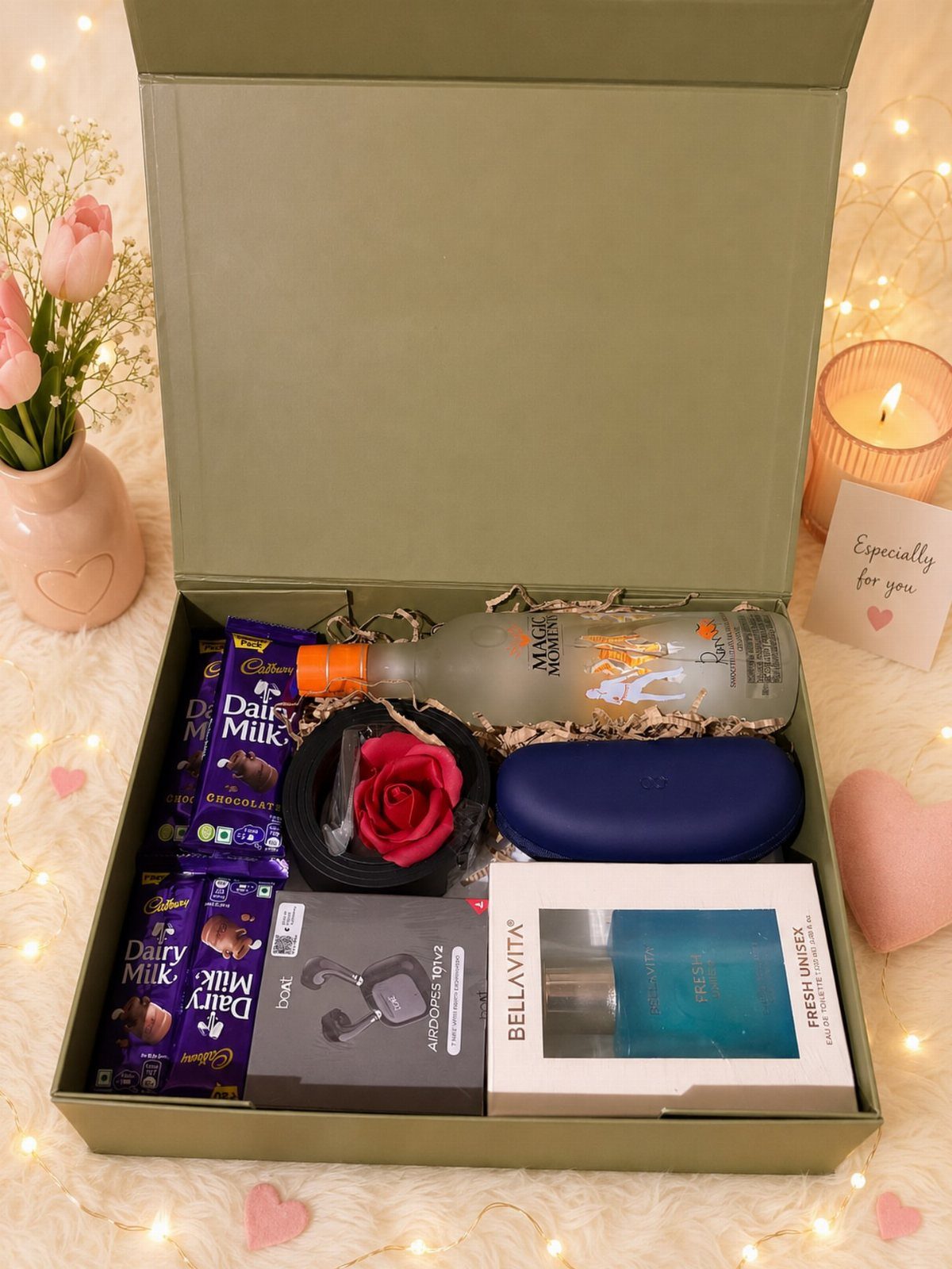 Gift box with chocolates, perfume, a red rose, and a bottle on fluffy white fabric.
