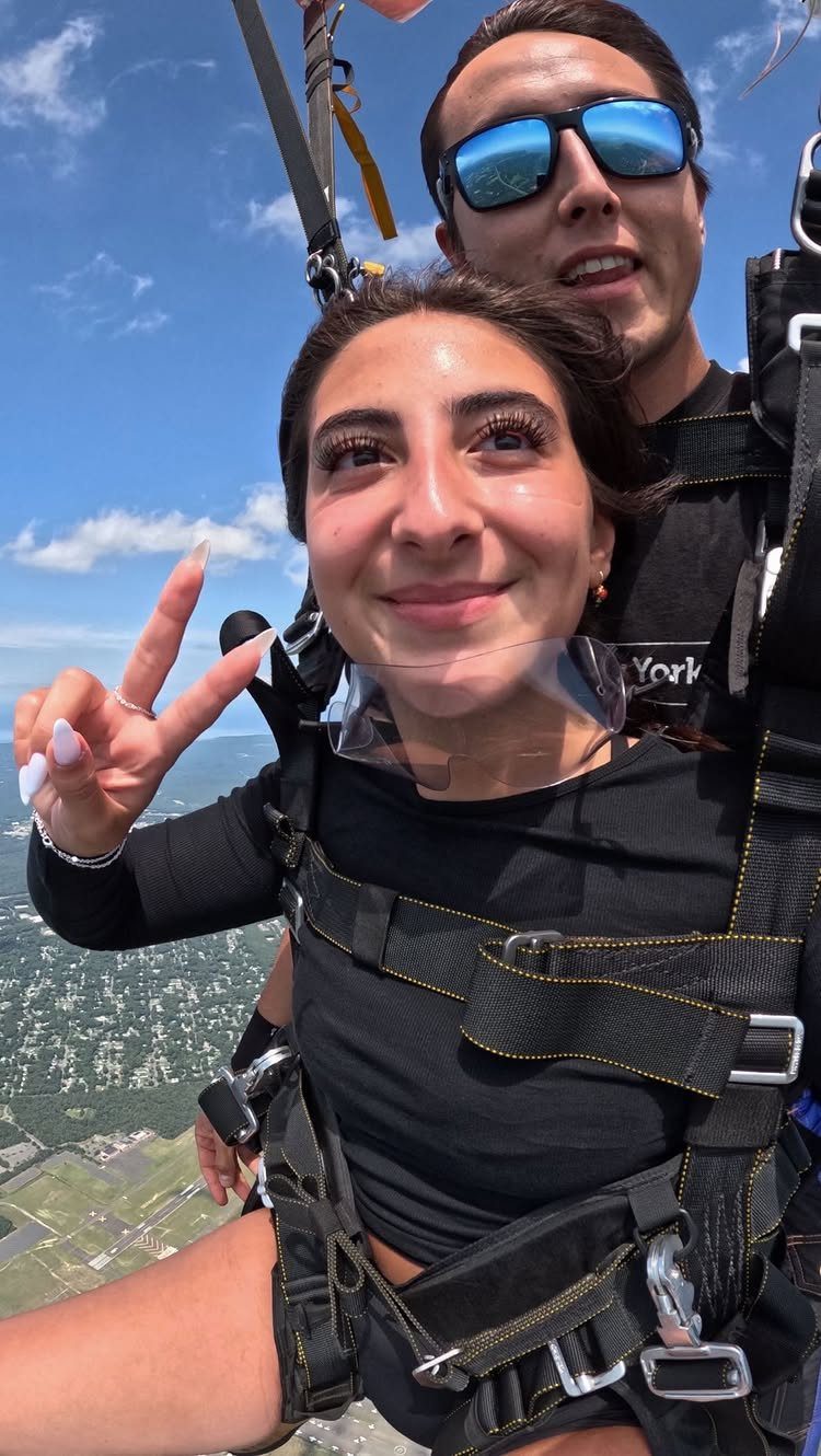 Woman and man skydiving together, smiling and making a peace sign in mid-air.
