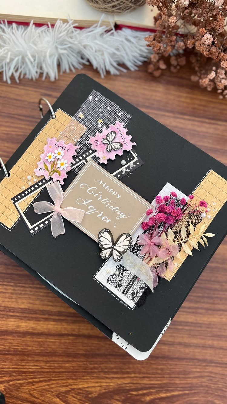 Black ring-bound scrapbook cover decorated with dried flowers, a butterfly, and birthday card.