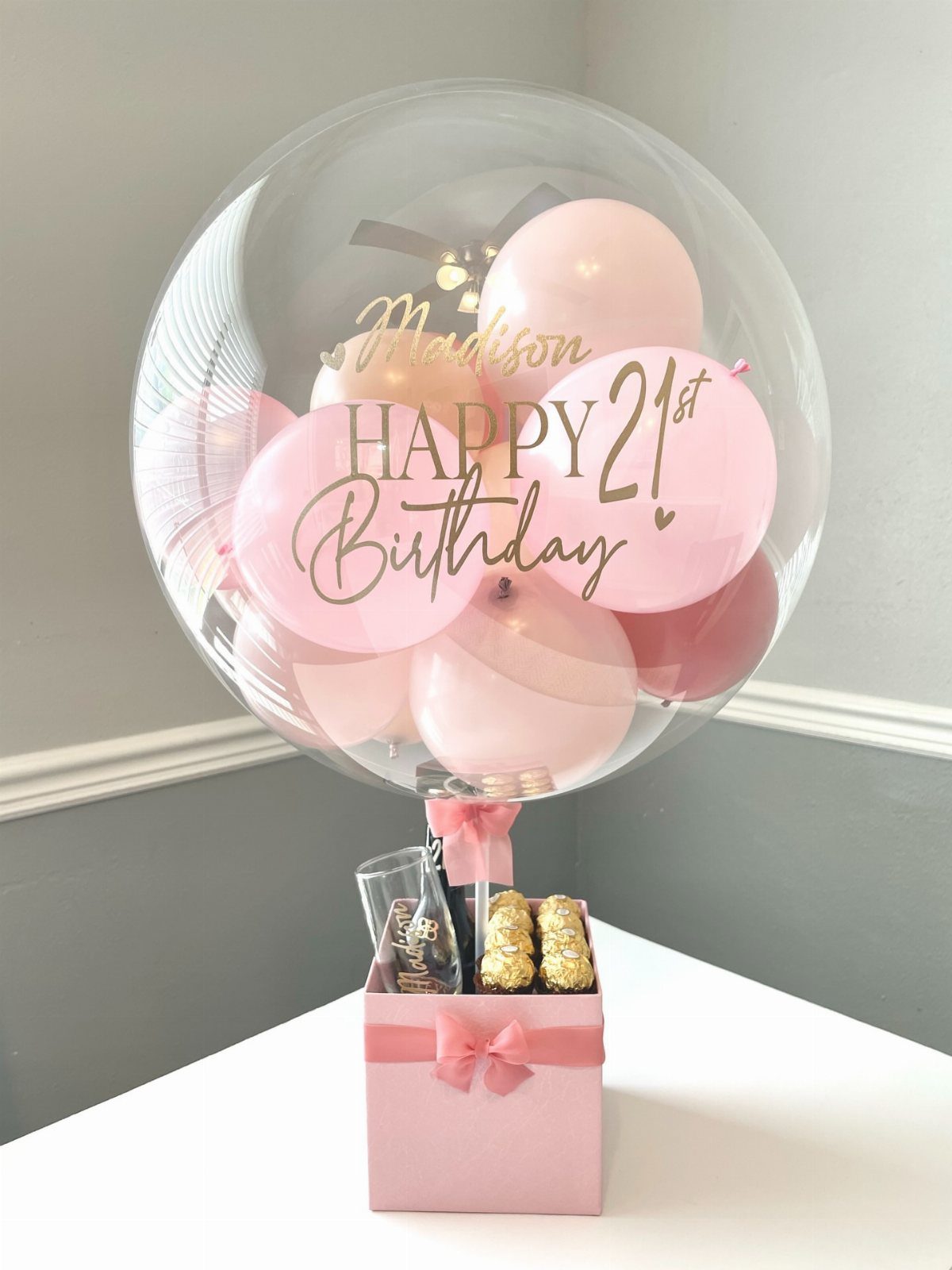 Large clear balloon with pink balloons inside and a pink gift box below.