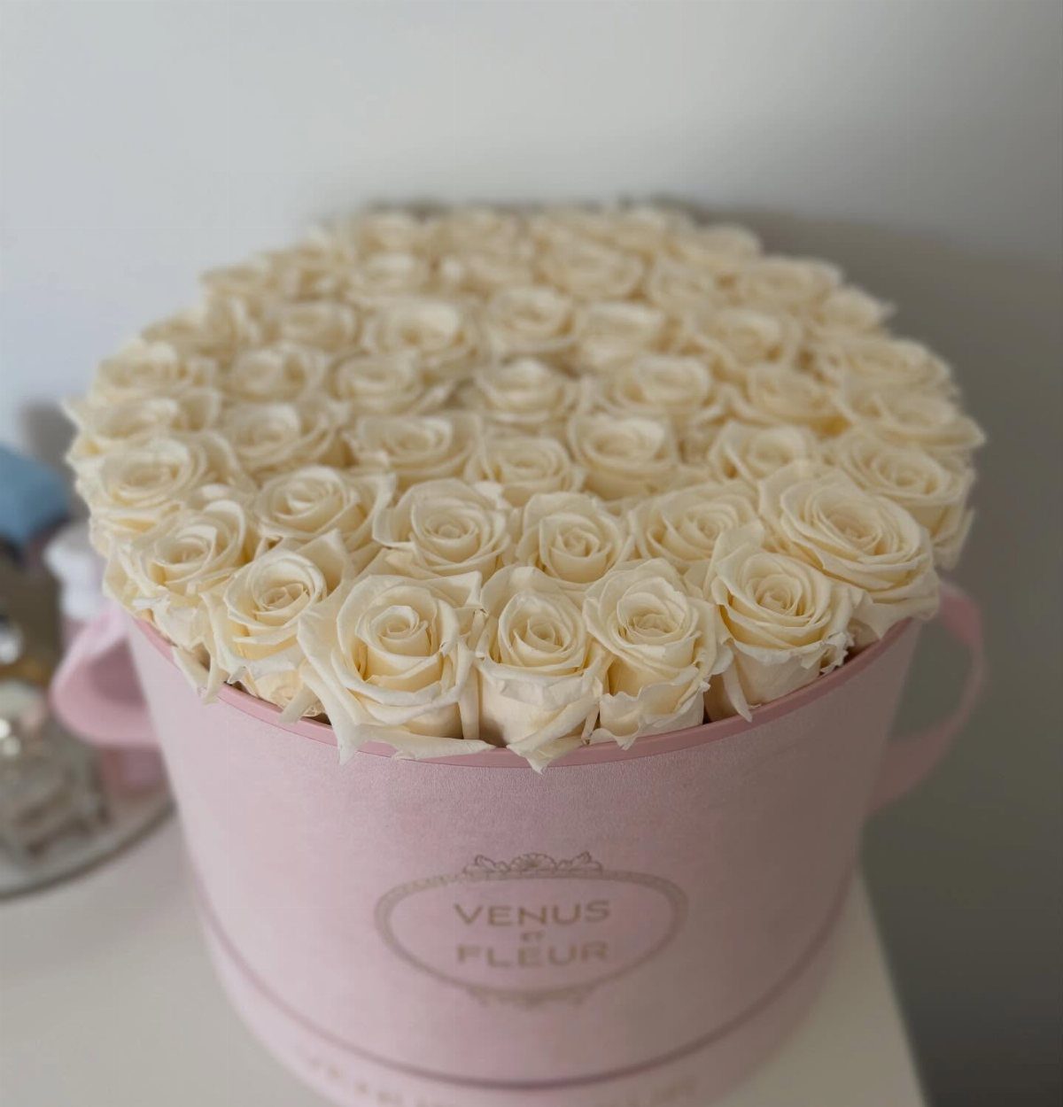 Cream-colored roses arranged in a round light pink hat box with a logo.