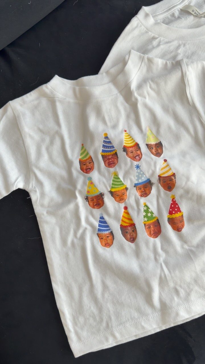 White t-shirt with a pattern of baby faces wearing colorful party hats.