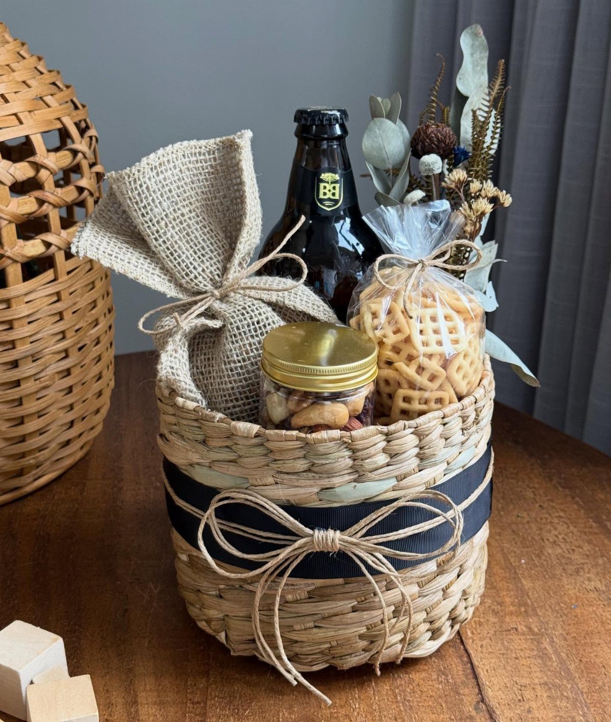 Woven gift basket with a beer bottle, snacks, and dried flowers on wood.