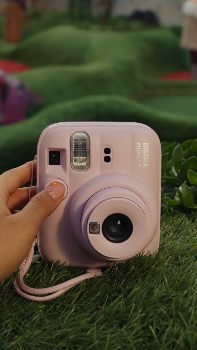 Light pink instant camera held by a hand on green grass.