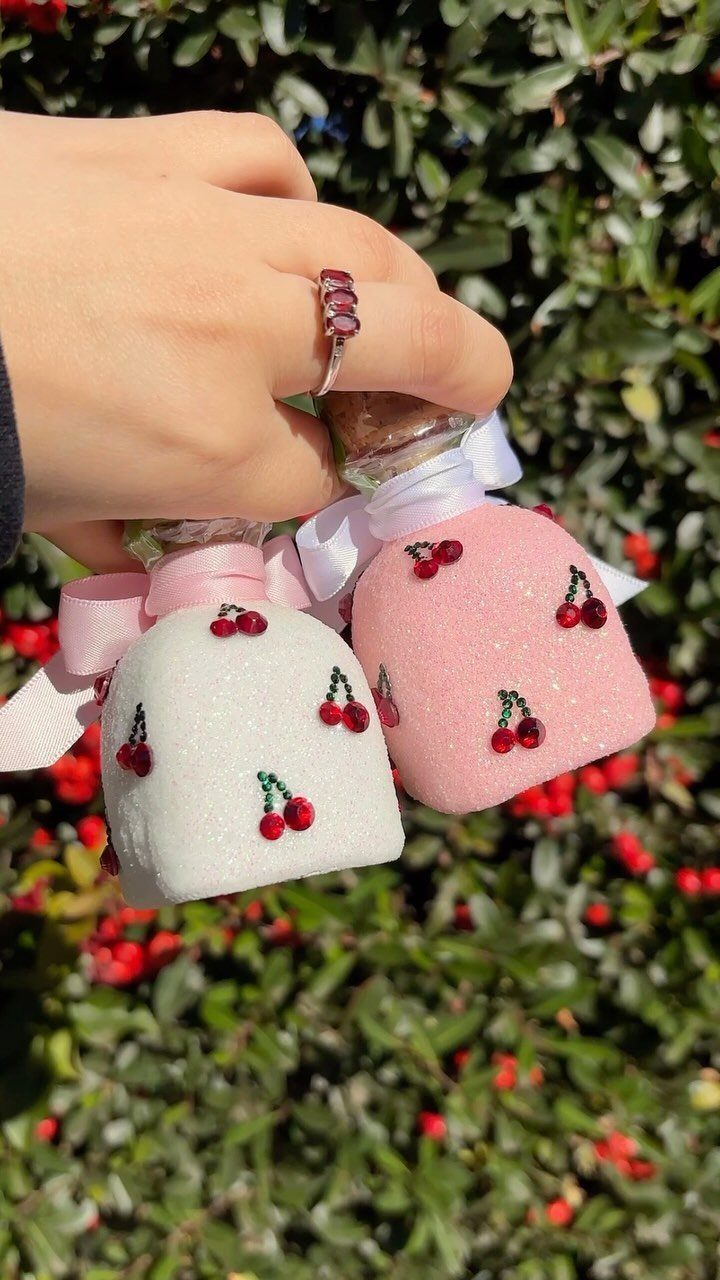 Two small glittery bottles decorated with cherries and ribbons held by a hand.