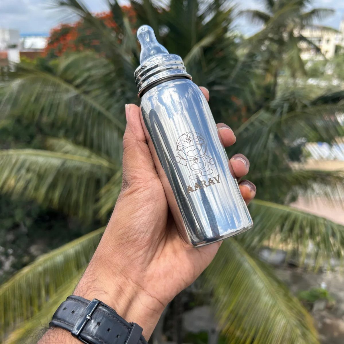 Silver metal baby bottle with engraved name held against palm tree background