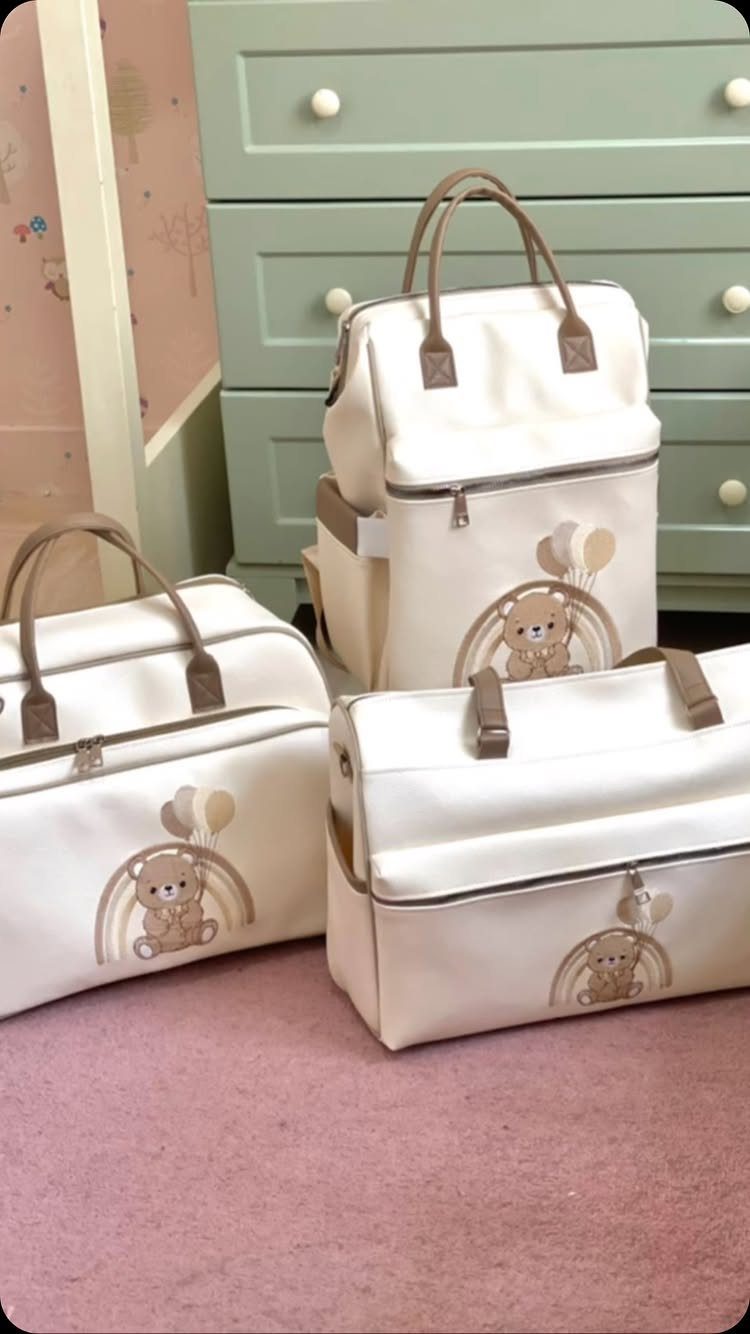 Three cream baby diaper bags with bear and rainbow designs on pink carpet.