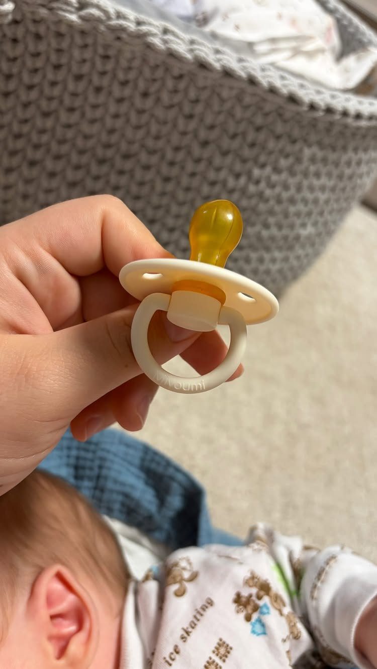 White and yellow pacifier held above a baby in a patterned onesie.