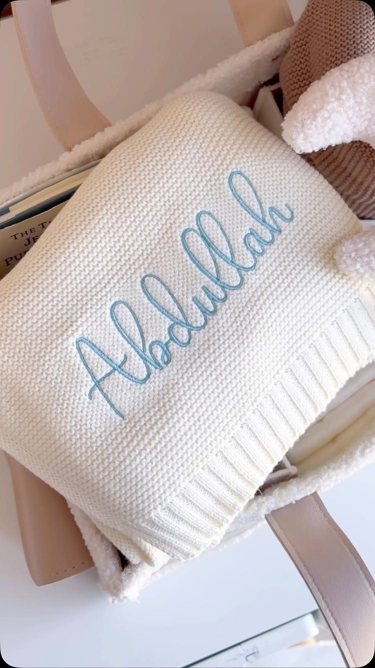 White knitted baby blanket with blue embroidered name in a fabric basket
