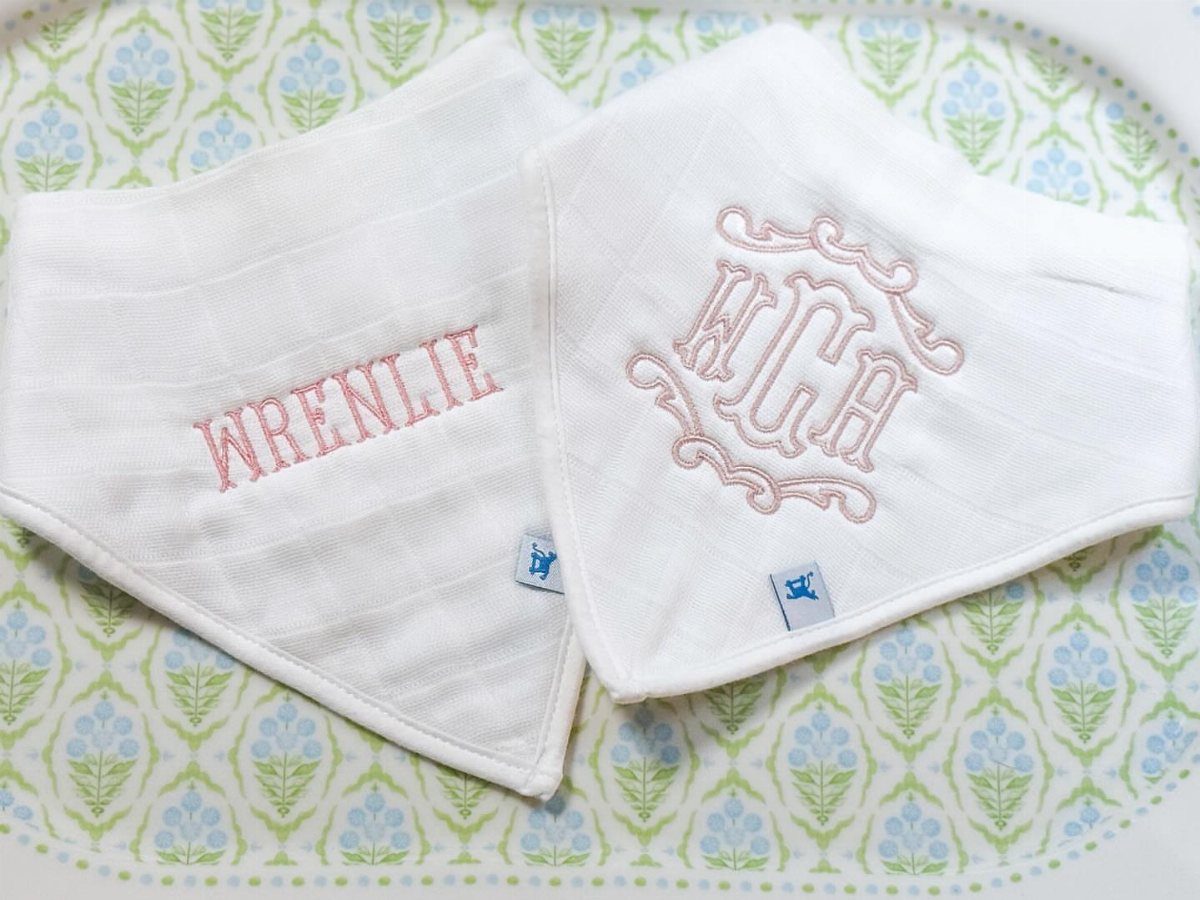 Two white embroidered baby bibs with pink lettering on a floral background