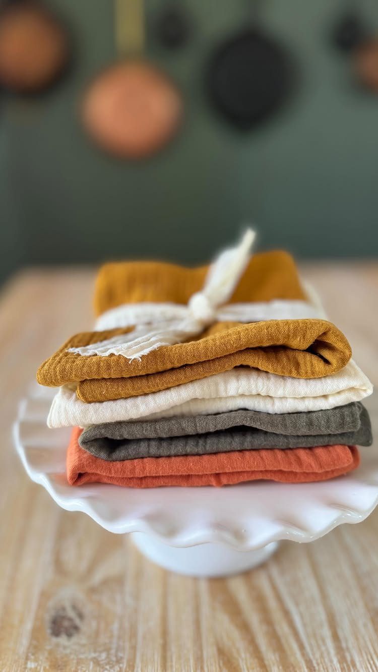 Stacked muslin baby cloths in earthy tones on a white scalloped cake stand.