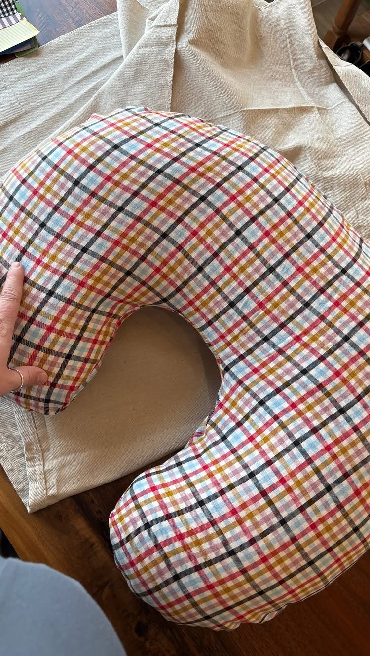 Plaid nursing pillow resting on a beige fabric surface with a hand touching it.