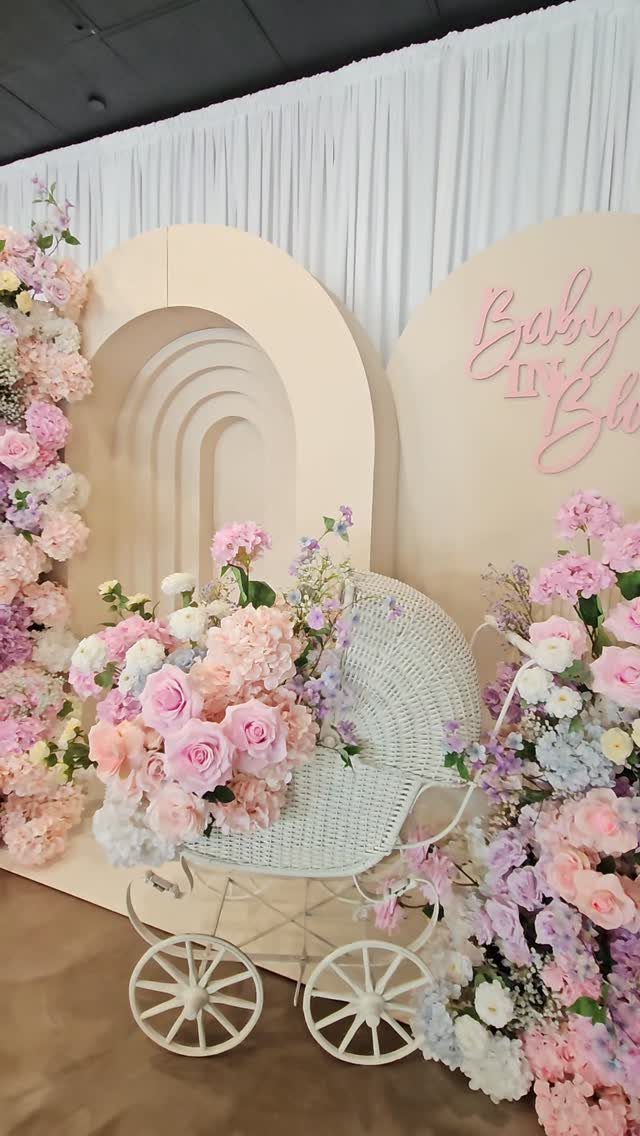 White wicker baby carriage filled with pastel flowers before a floral arched backdrop.
