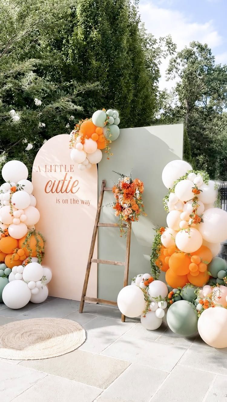 Peach arched backdrop with orange and white balloons and a wooden flower ladder.
