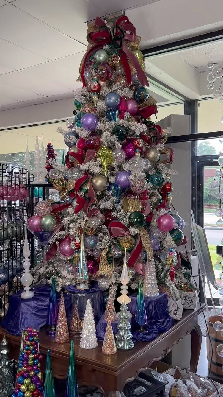 Frosted Christmas tree decorated with rich jewel-toned ornaments and red ribbons on a table.