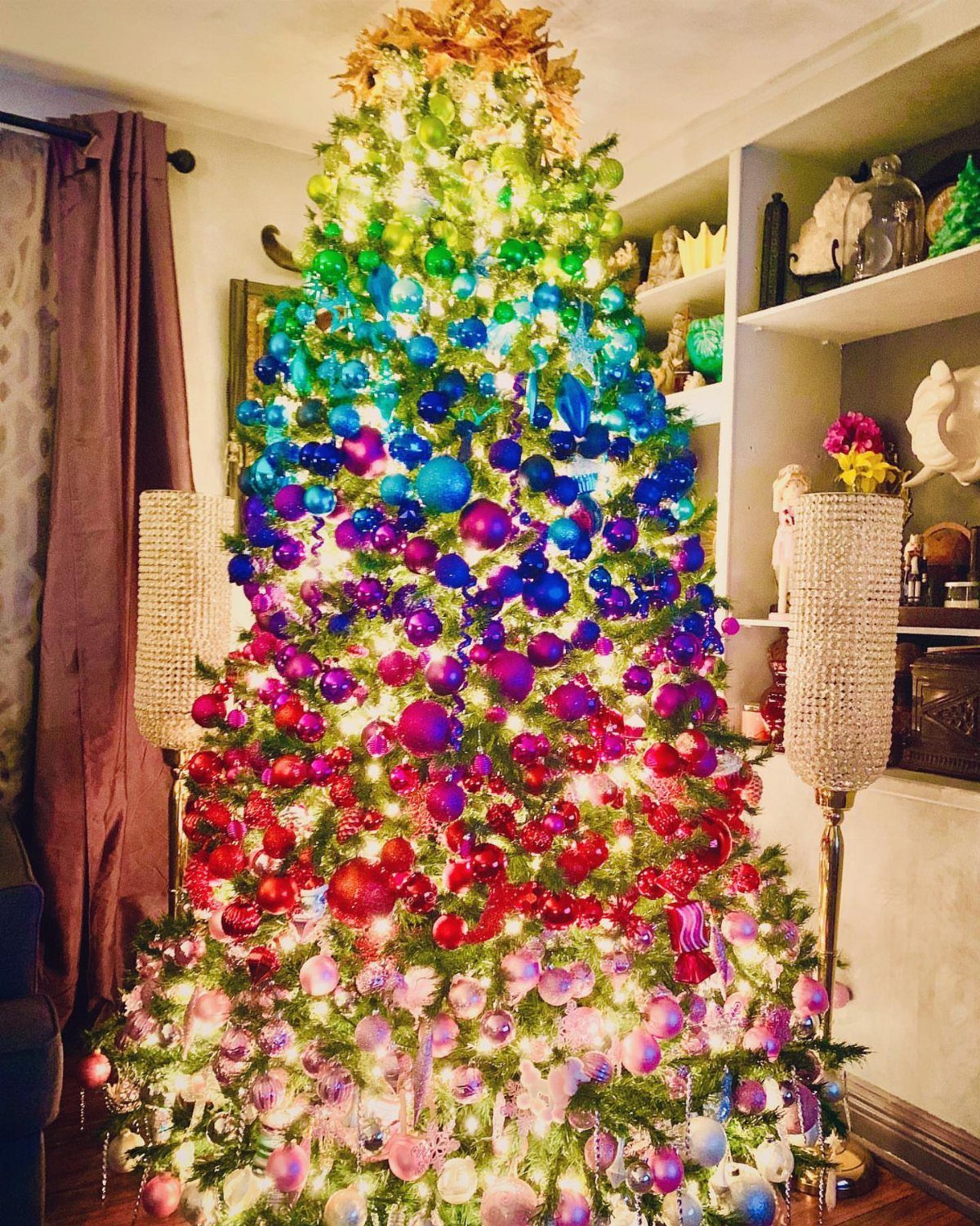 Rainbow ombre Christmas tree with colorful ornaments and bright lights in a room.