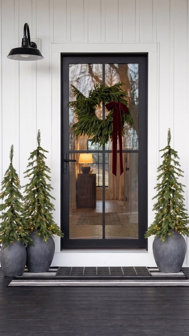 Three small lit Christmas trees in grey pots flanking a decorated front door.