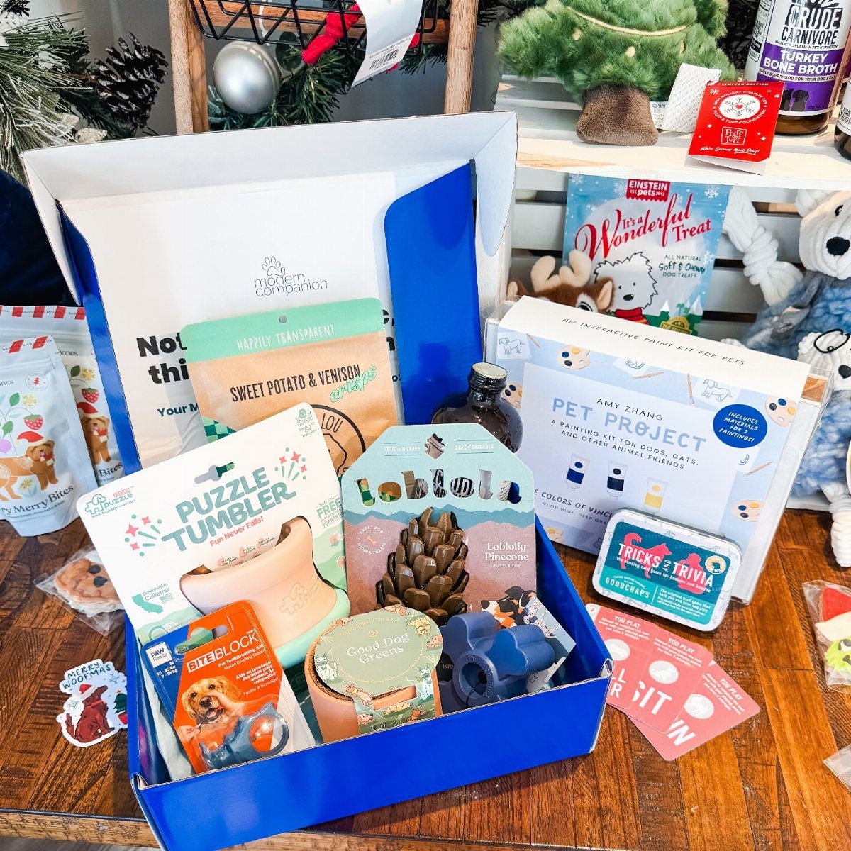 Dog gift box filled with treats and toys next to a small Christmas tree.