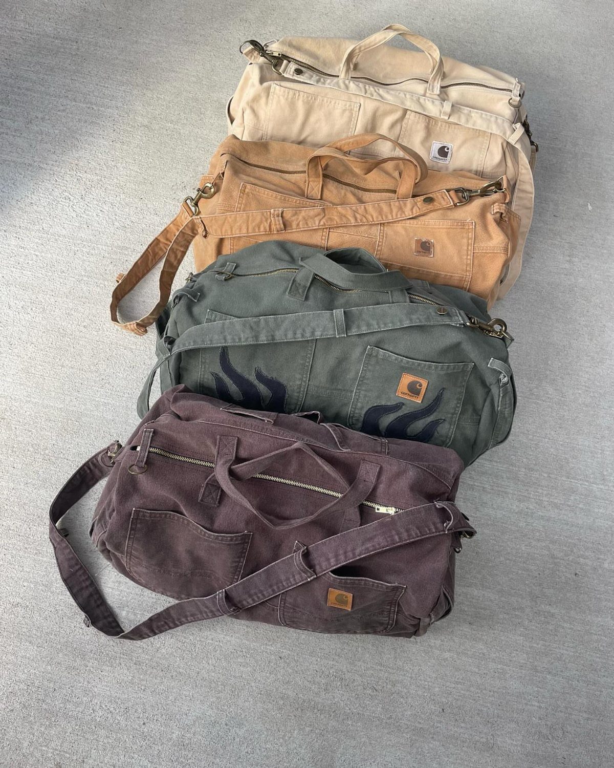 Four Carhartt canvas duffel bags in different colors stacked on a concrete floor.