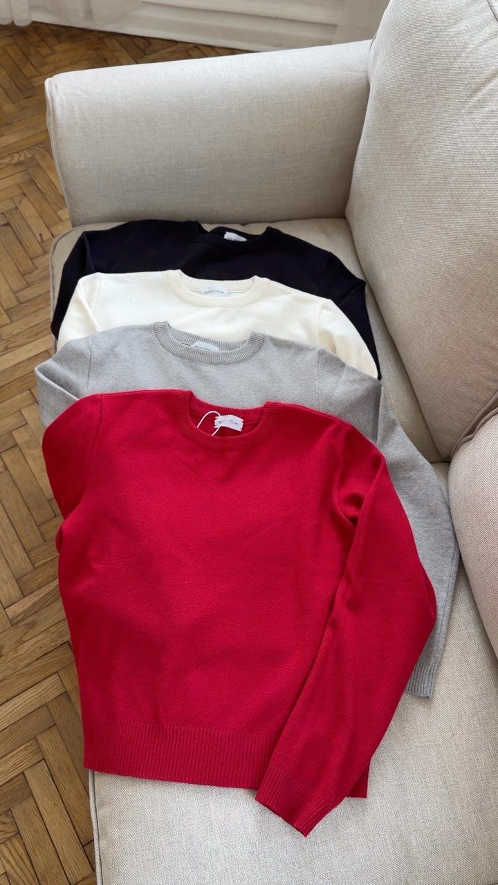 Four colorful sweaters stacked on a beige sofa over a wooden floor.