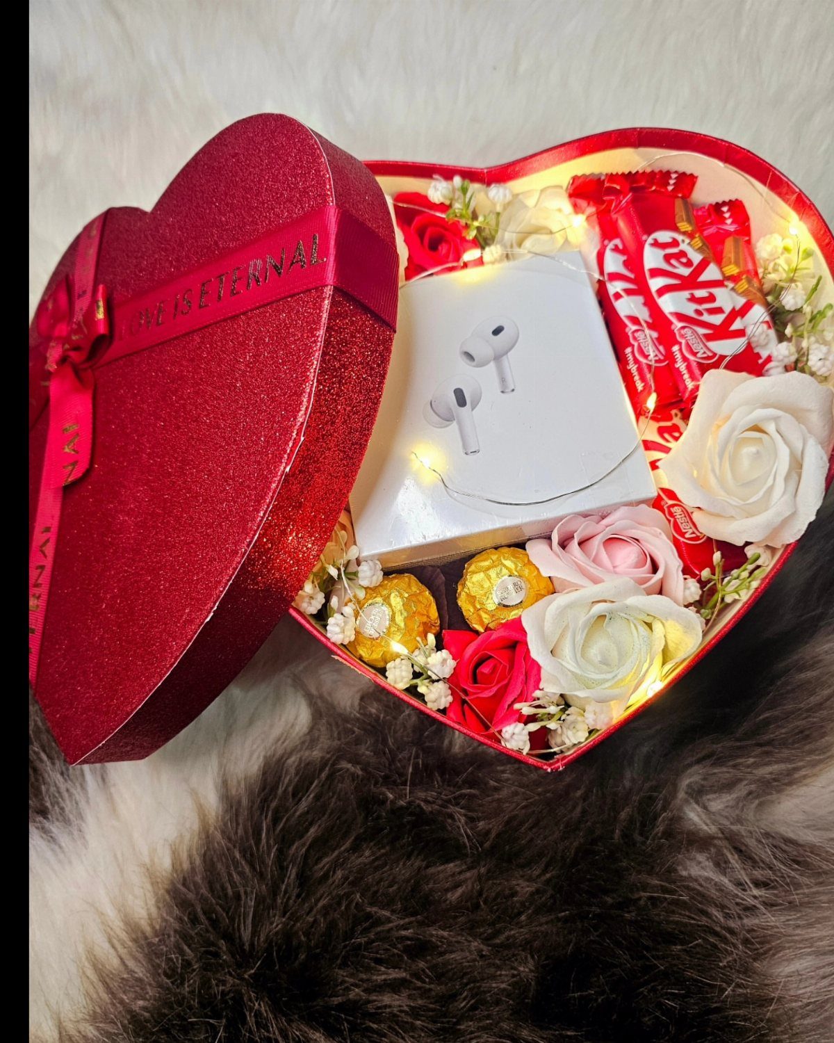 Heart-shaped red gift box filled with earbuds, chocolates, and artificial roses on fur.