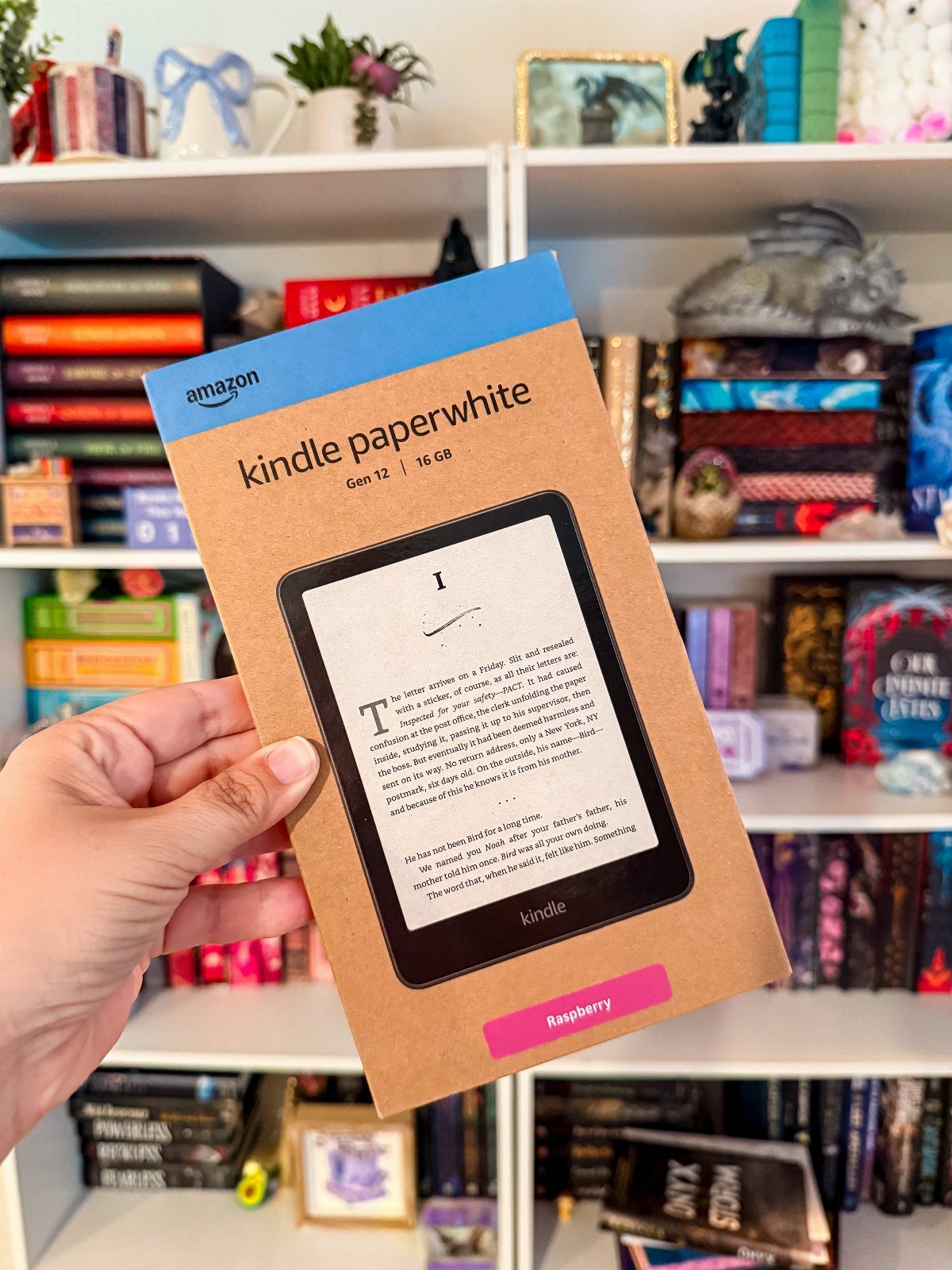Kindle Paperwhite packaging held in front of a bookshelf filled with colorful books.