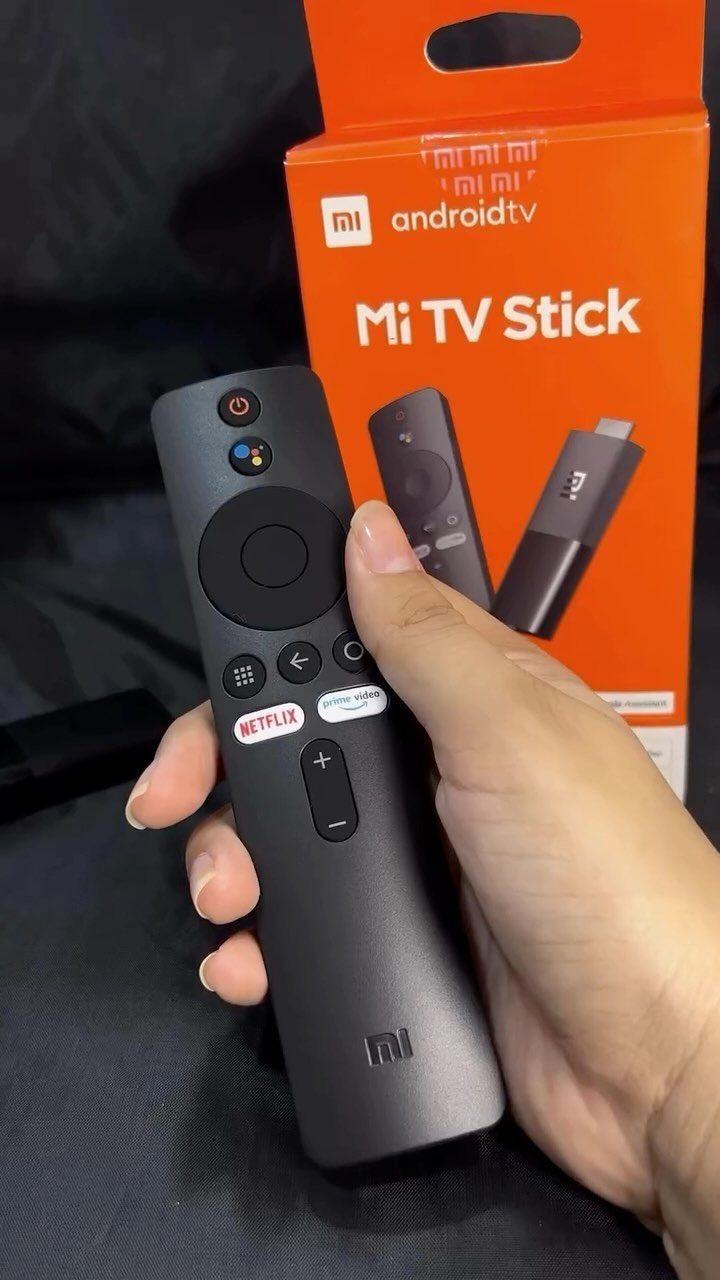 Hand holding a black Mi TV Stick remote with orange product packaging behind it.