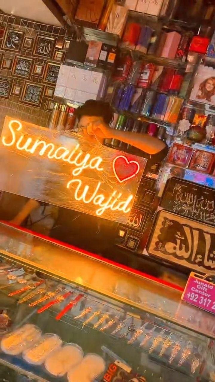 Glowing orange neon sign with names and a heart held in a shop.
