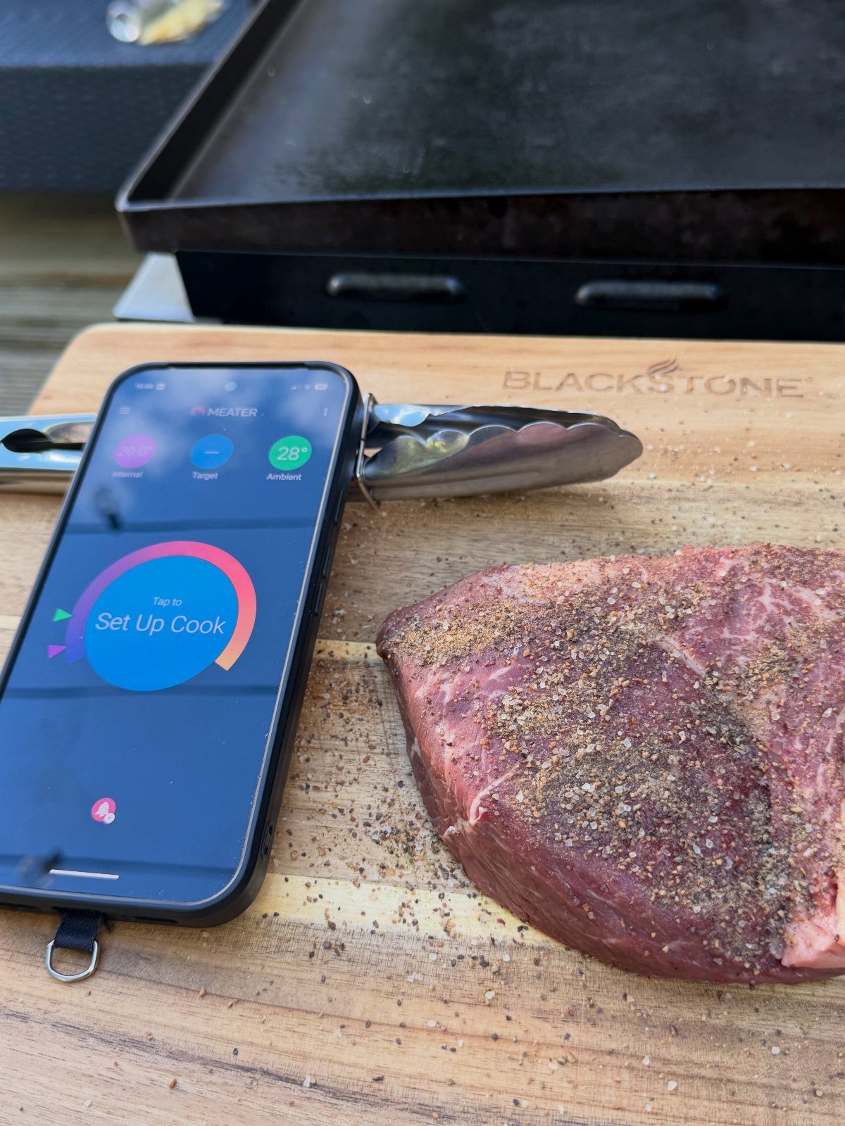 Smartphone with cooking app next to seasoned raw steak on wooden cutting board