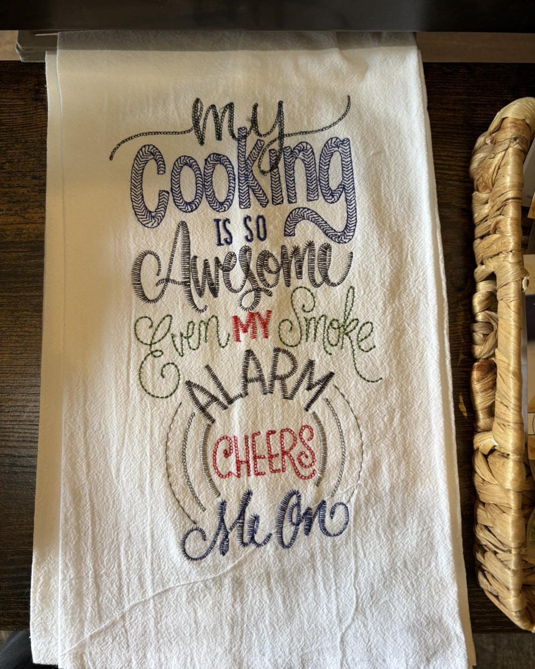 White kitchen towel with humorous embroidered text about cooking and smoke alarms.