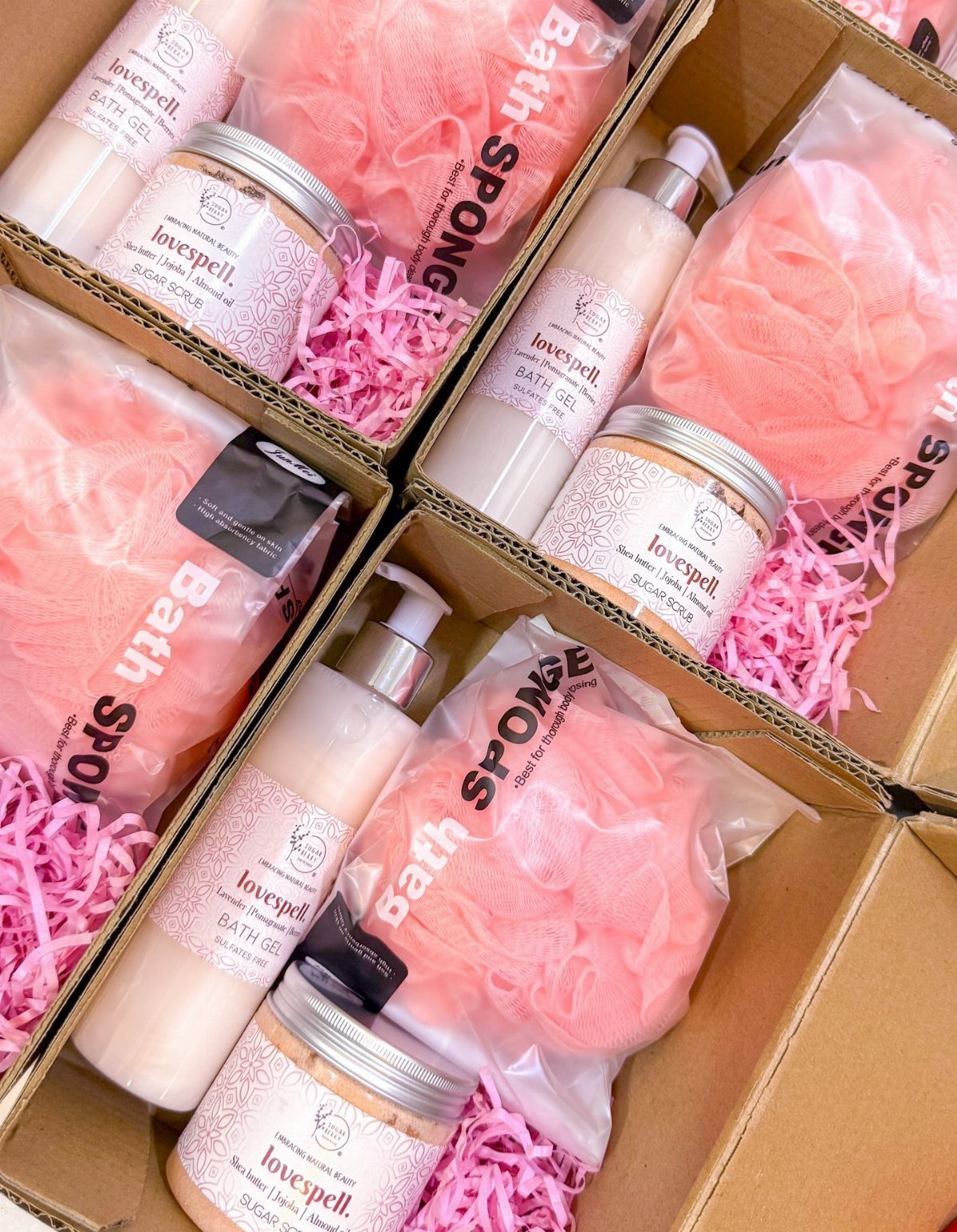 Four gift boxes containing pink bath sponges, bath gel, and sugar scrub jars.
