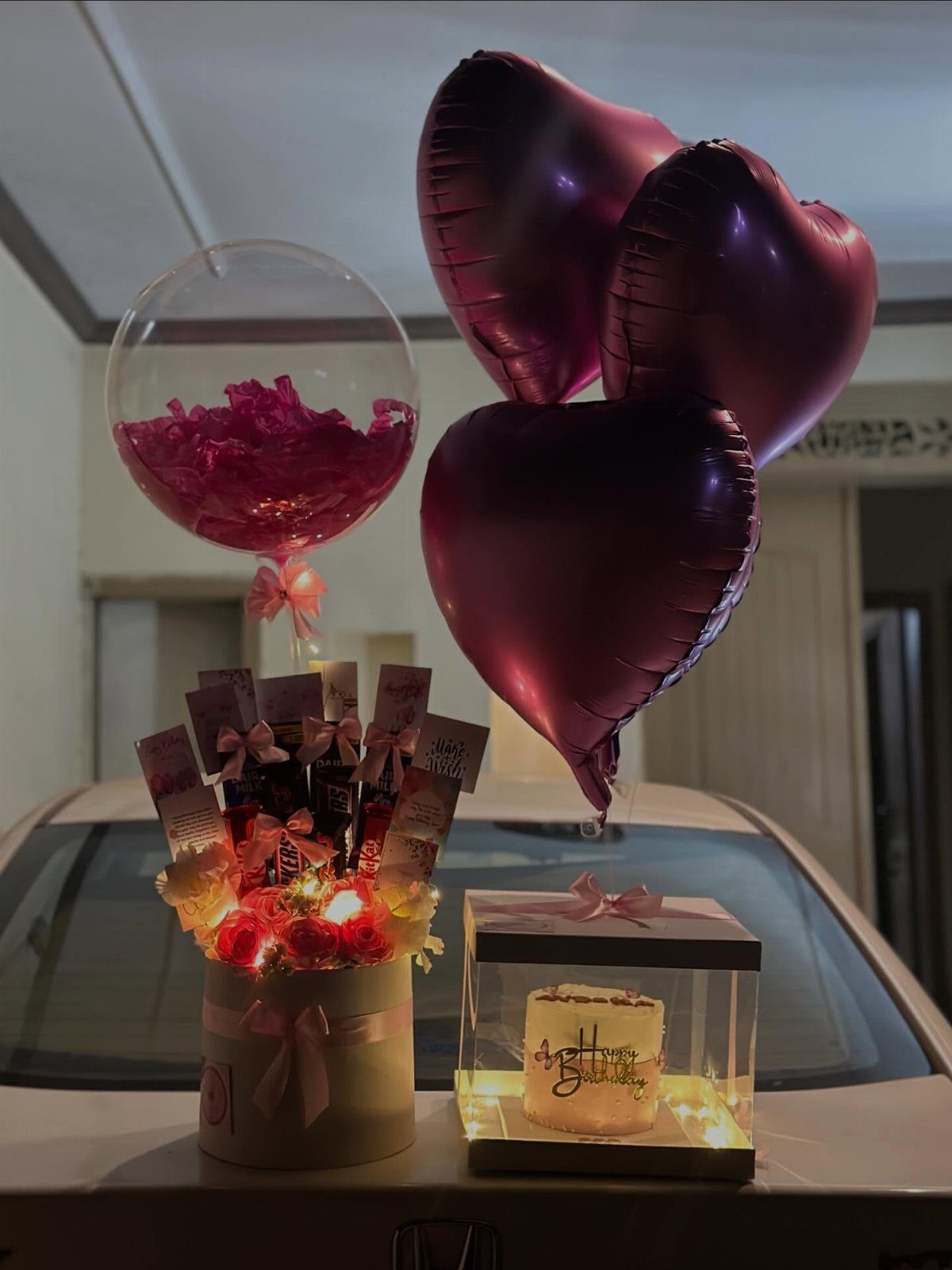 Gift arrangement with purple heart balloons, flowers, chocolates, and a small cake.