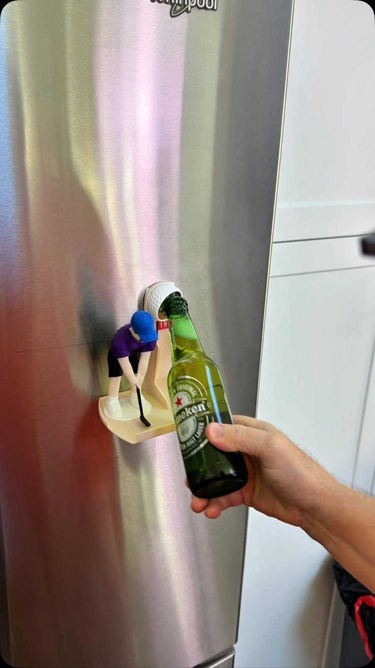 Hand opening a beer bottle using a golfer-shaped wall-mounted bottle opener on a fridge.