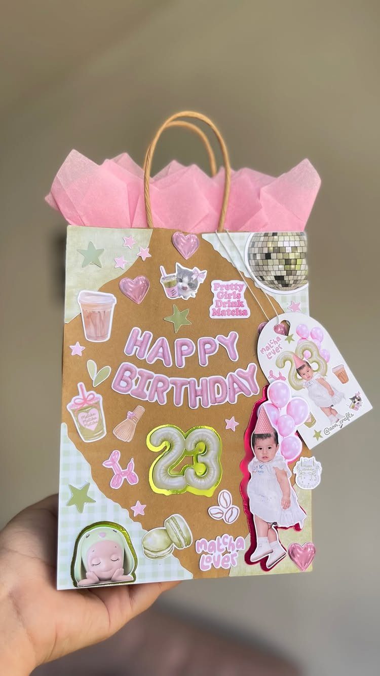 Decorated birthday gift bag with pink tissue paper, stickers, and baby photos.
