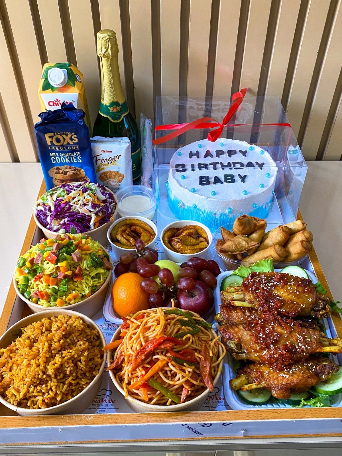 Birthday feast tray with blue cake, champagne, noodles, chicken, and various side dishes.