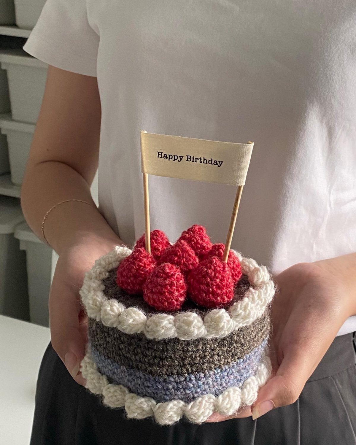Crochet birthday cake with red strawberries and a Happy Birthday sign held by hands