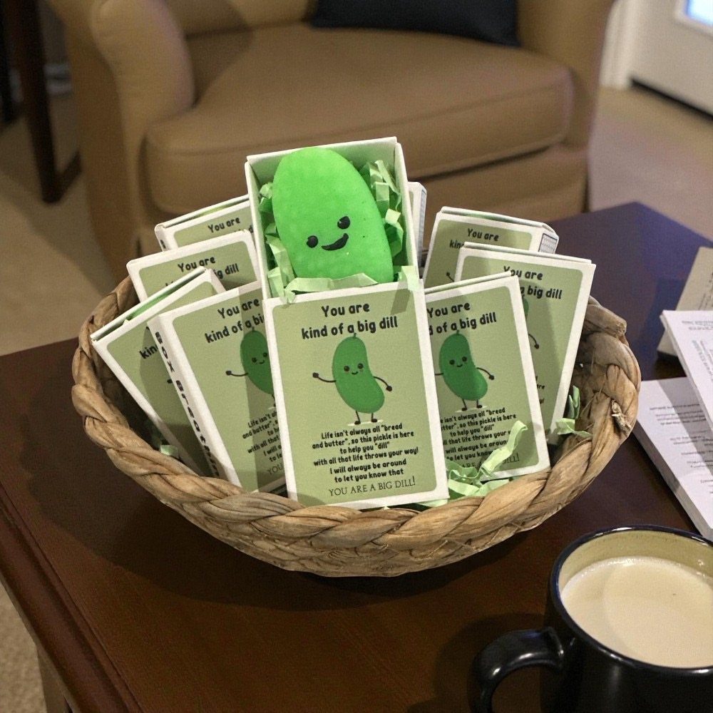 Plush green pickles in gift boxes inside a woven basket on a table.