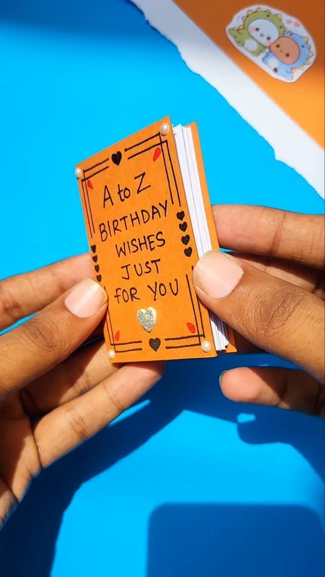 Small handmade orange birthday wishes notebook held by two hands over blue background