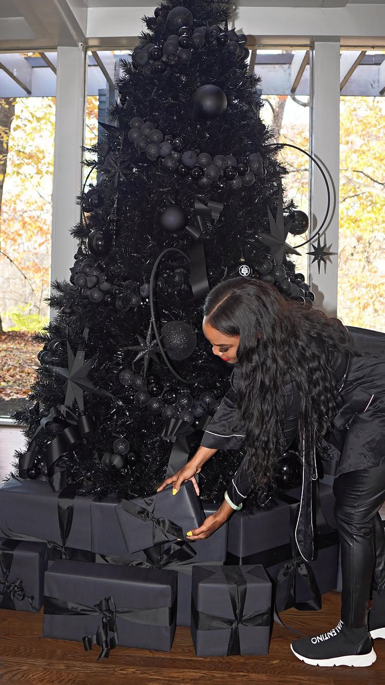 Woman standing by a black Christmas tree decorated with black ornaments and gifts.