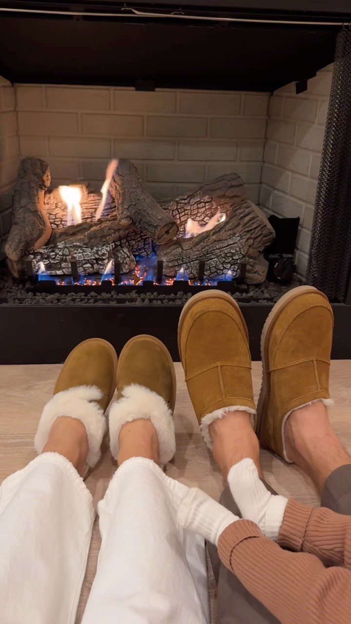 Two pairs of tan suede slippers with white fur lining by a fireplace.