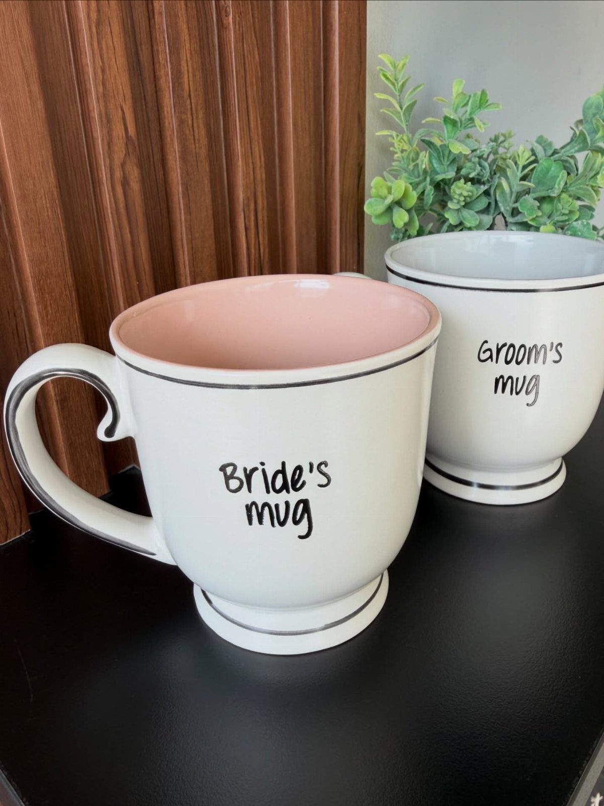 Two white ceramic mugs labeled Bride's mug and Groom's mug on a black table.