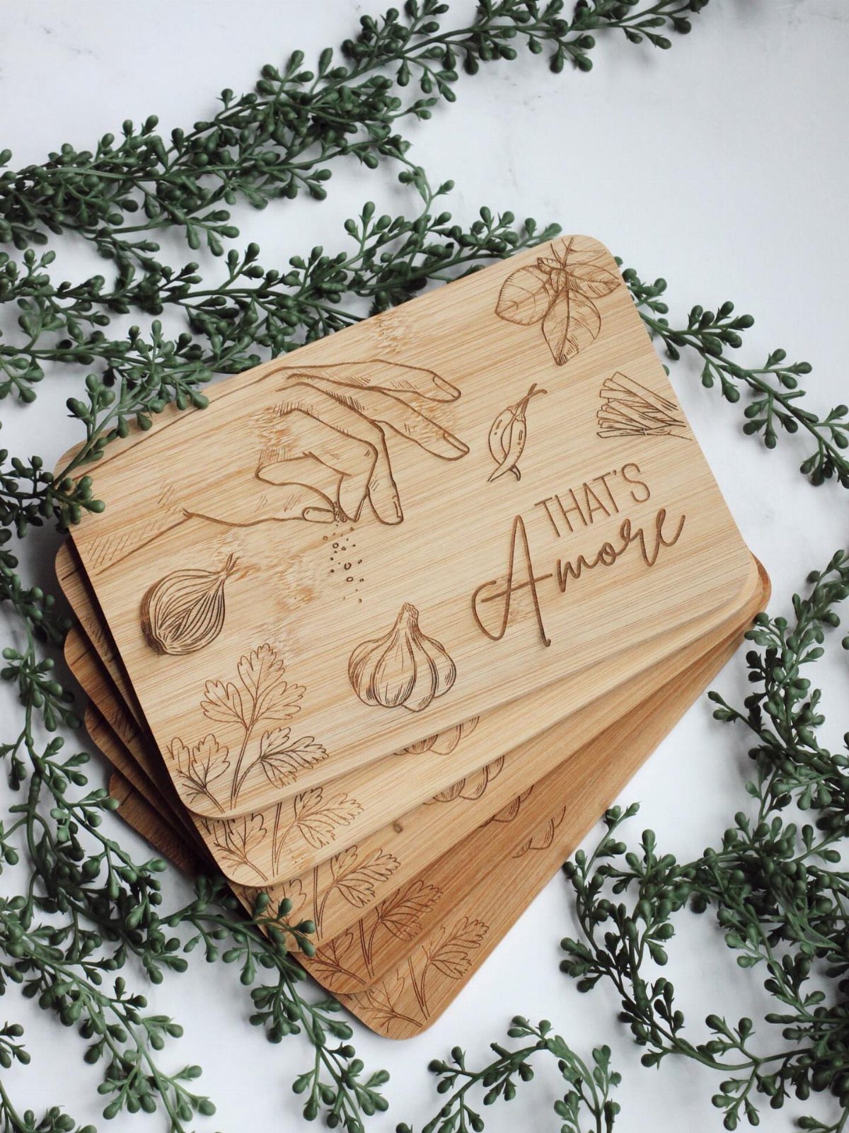 Engraved wooden cutting boards with garlic illustrations and That's Amore text among greenery