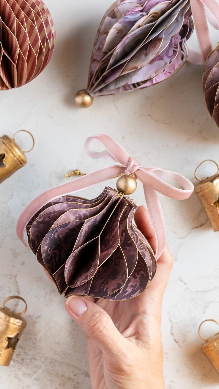 Purple honeycomb paper ornament held by a hand with pink ribbon and gold bells.