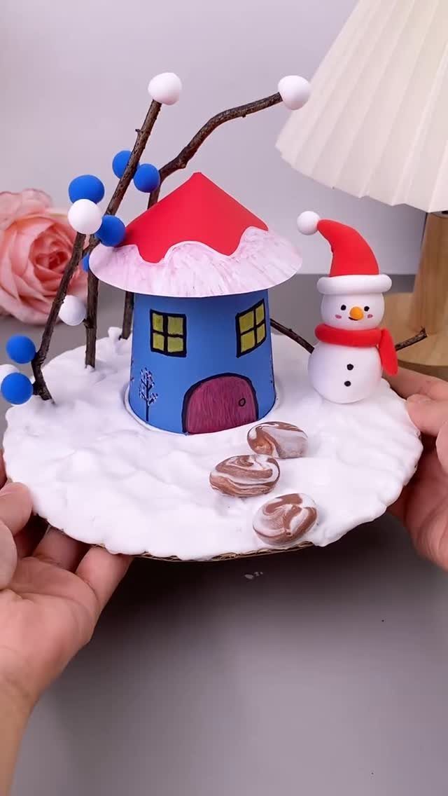 Small blue paper house with red roof and snowman on snowy base.
