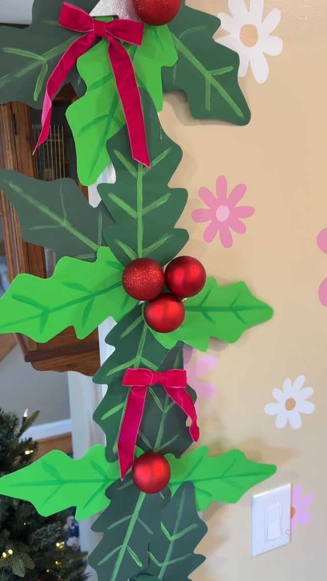Paper holly leaves and red berries with ribbons decorating a beige wall