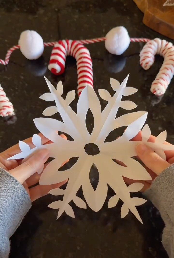 White paper snowflake held by hands with a candy cane garland behind it