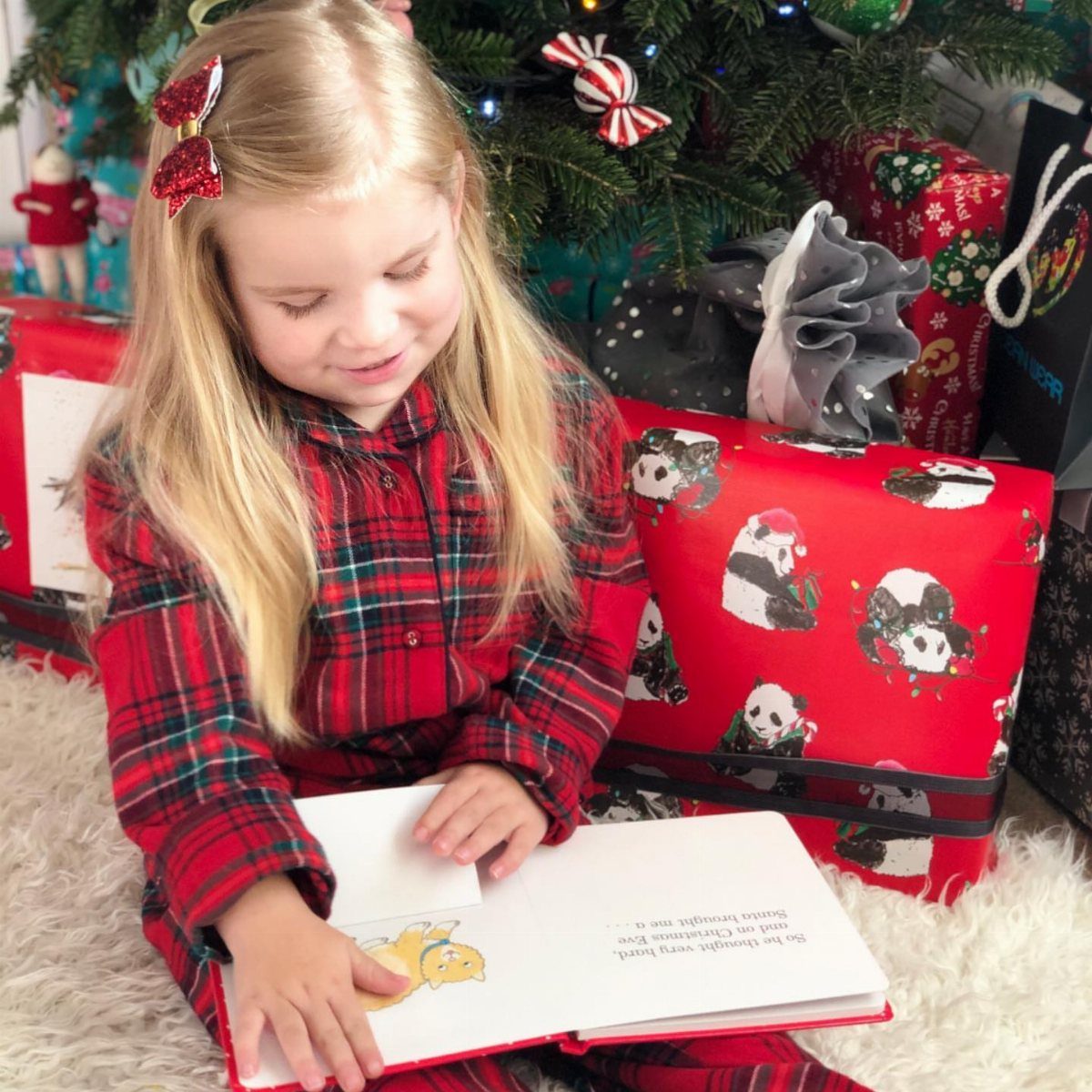 Young girl in red plaid pajamas reading a Christmas book among wrapped presents.