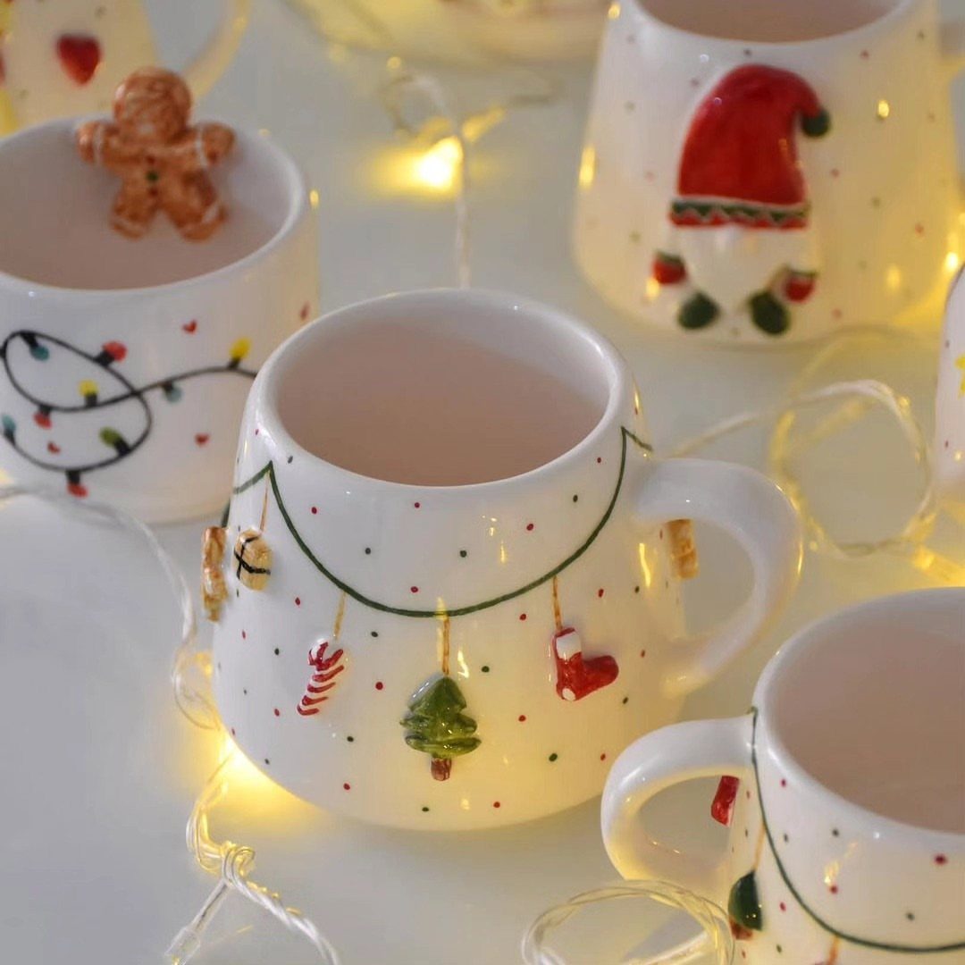 White ceramic mugs with Christmas decorations and warm fairy lights on white surface
