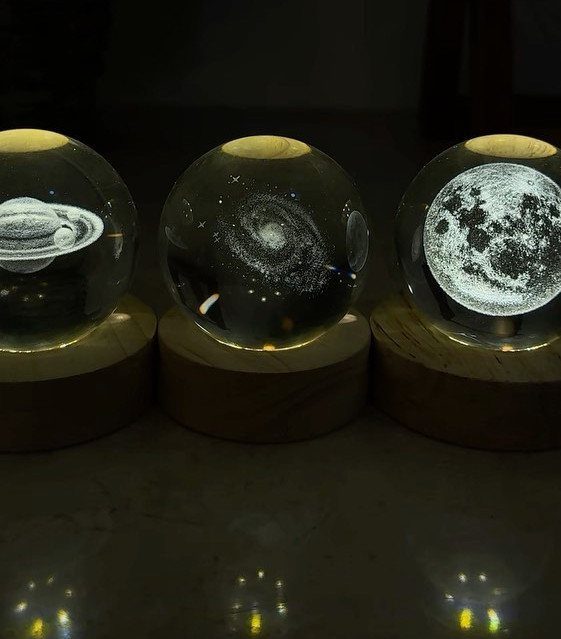 Three glowing glass spheres with laser-etched space scenes on wooden bases.